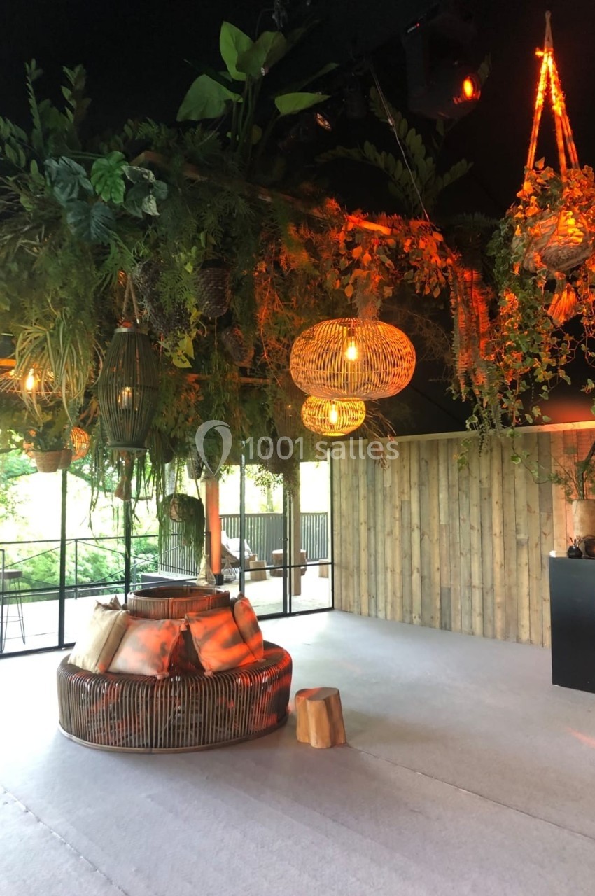 Espace intérieur décoré avec des plantes suspendues, des luminaires en rotin et un foyer central entouré de sièges.