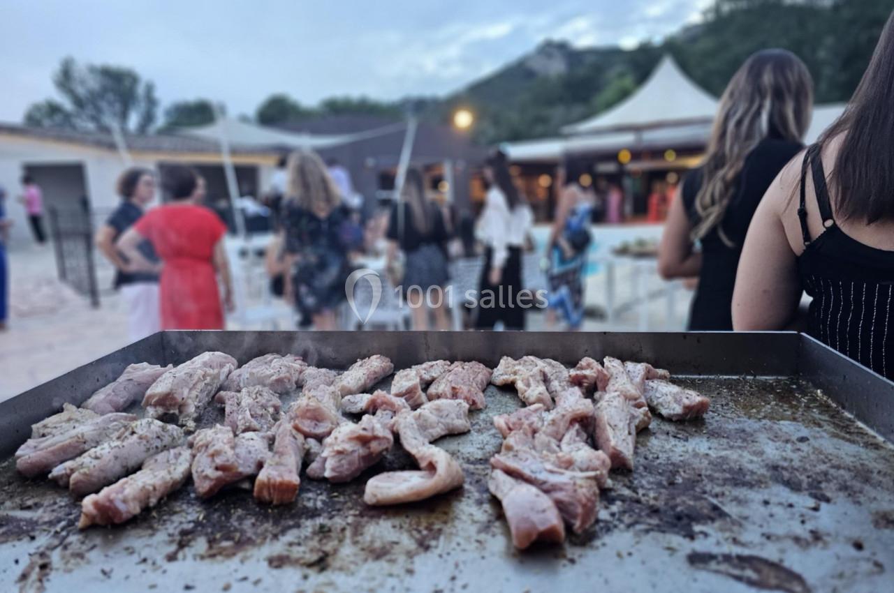 Viande en train de griller sur une plancha lors d'un événement en plein air avec des invités en arrière-plan.