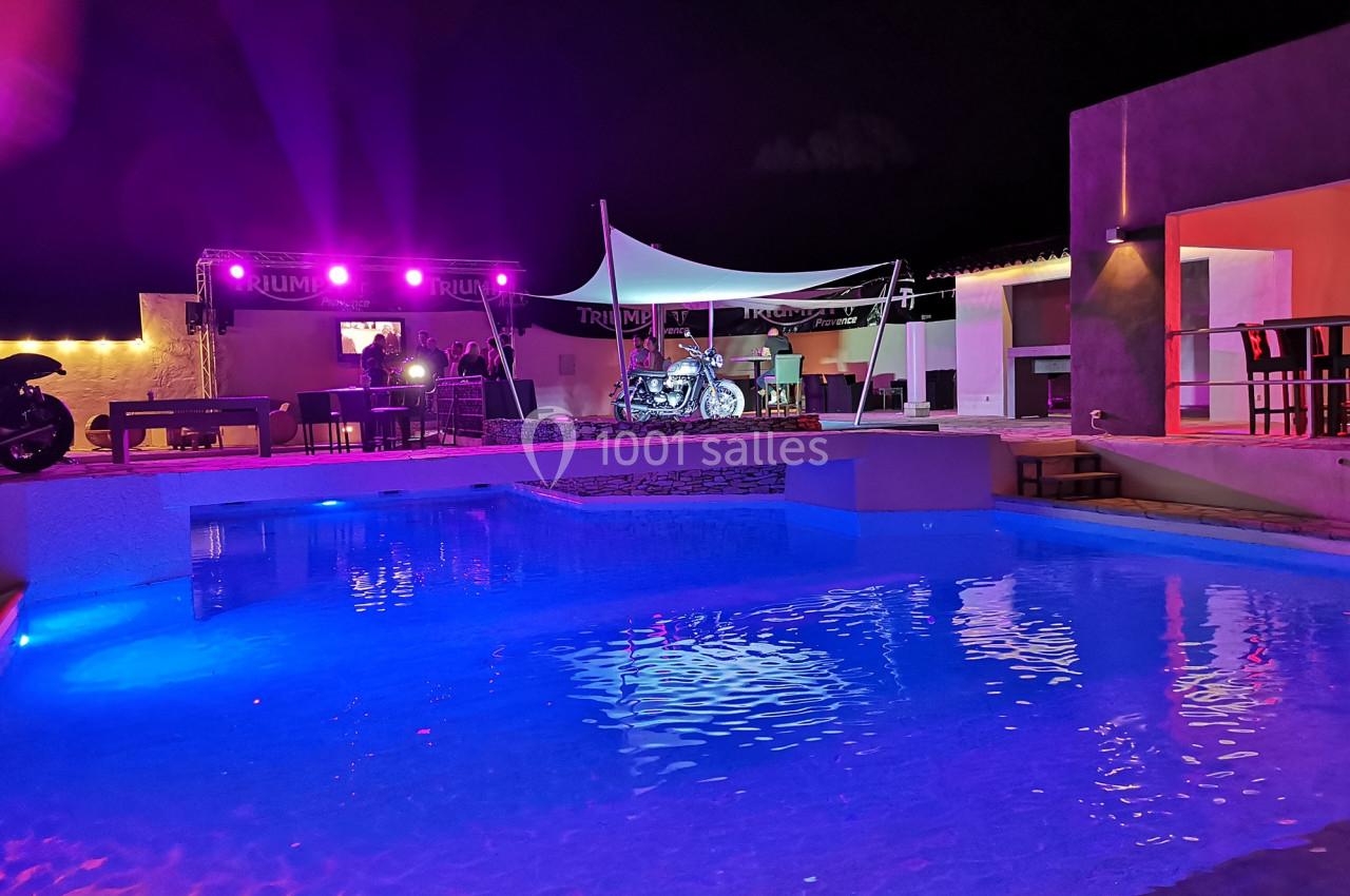 Piscine éclairée de nuit avec une terrasse animée en arrière-plan, incluant des lumières colorées et une moto exposée.