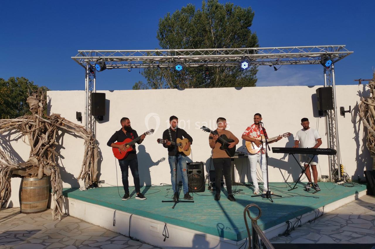 Un groupe de musiciens joue sur une petite scène extérieure avec guitares, clavier et éclairage scénique.