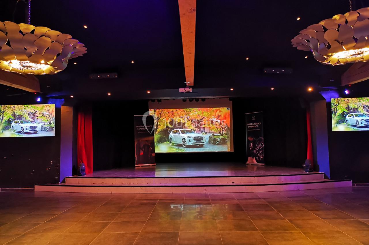 Scène éclairée avec écran central affichant une voiture blanche devant un paysage automnal, entourée de projecteurs.