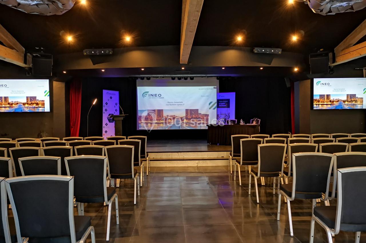 Salle de conférence vide avec des rangées de chaises, une scène équipée d'écrans et un éclairage tamisé.