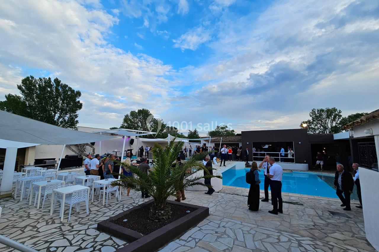 Personnes rassemblées autour d'une piscine dans un espace extérieur avec mobilier blanc et ciel partiellement nuageux.
