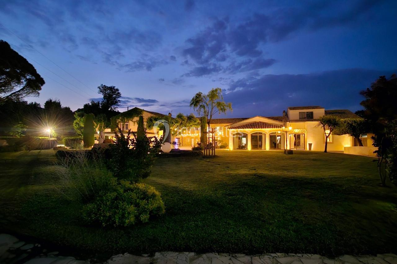 Vue nocturne d'un grand jardin éclairé avec une maison aux façades illuminées et un ciel crépusculaire en arrière-plan.