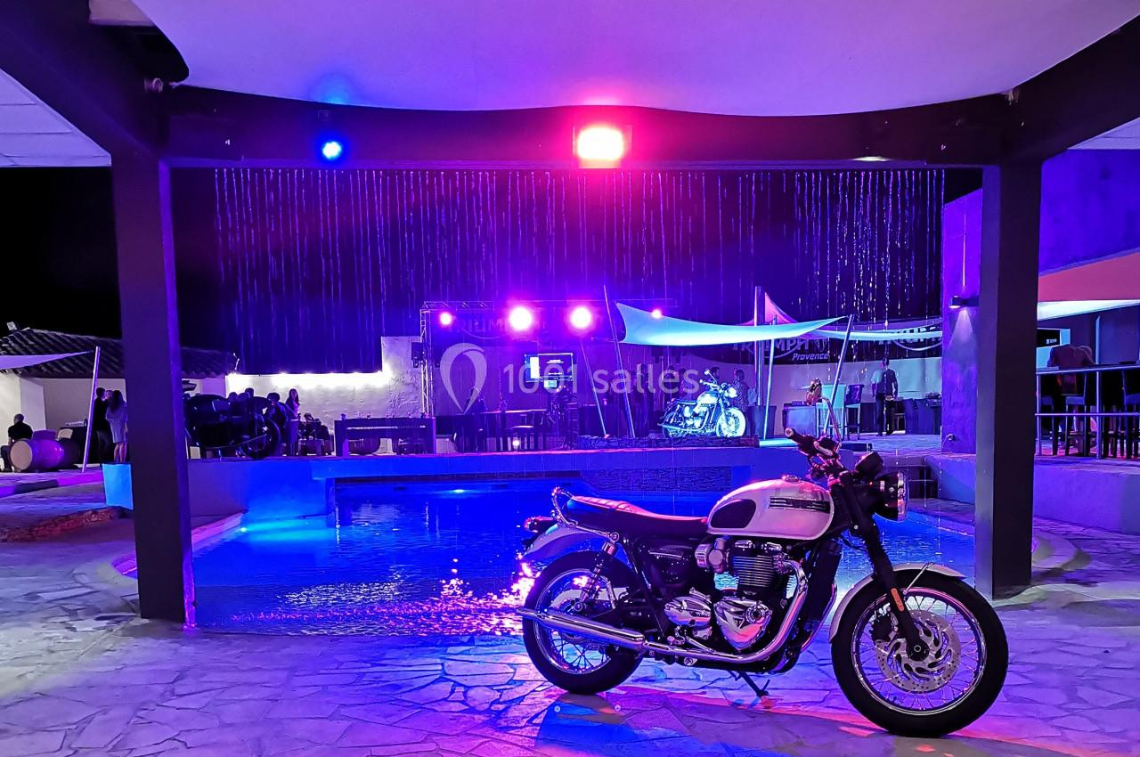 Une moto blanche et noire stationnée devant une piscine éclairée par des lumières colorées dans un cadre nocturne.