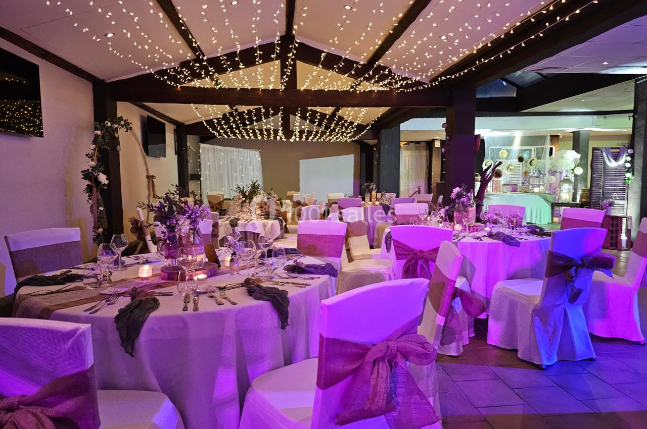 Salle de réception décorée pour un mariage, avec tables dressées, chaises ornées de nœuds et guirlandes lumineuses…