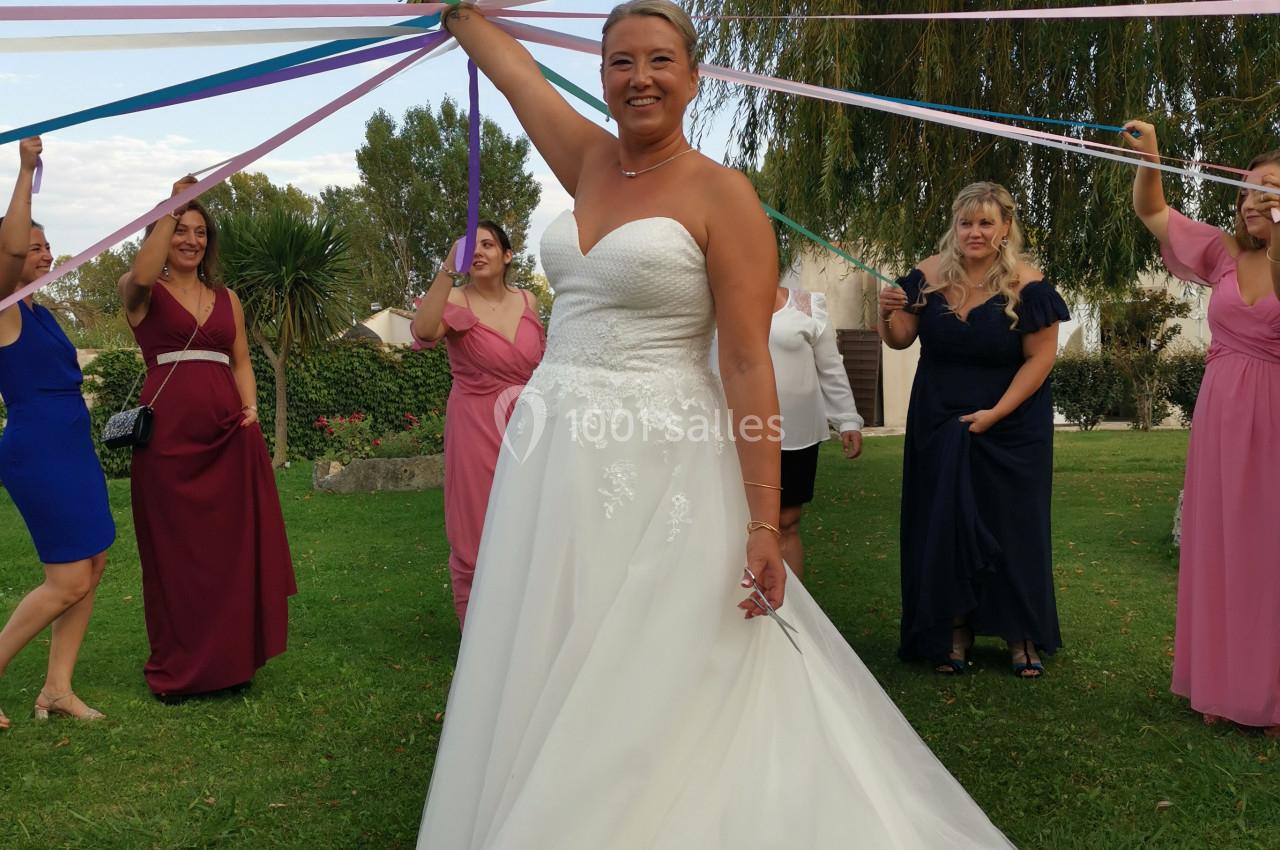 Une mariée en robe blanche tient un bouquet sous des rubans colorés, entourée d'invitées dans un jardin.
