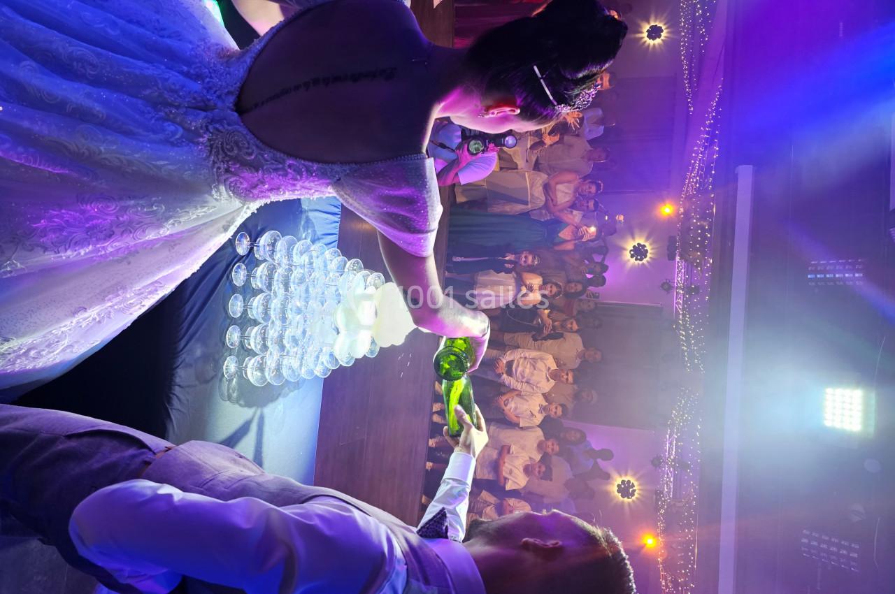 Un couple remplit une pyramide de verres avec du champagne lors d'une réception festive éclairée de lumières colorées.