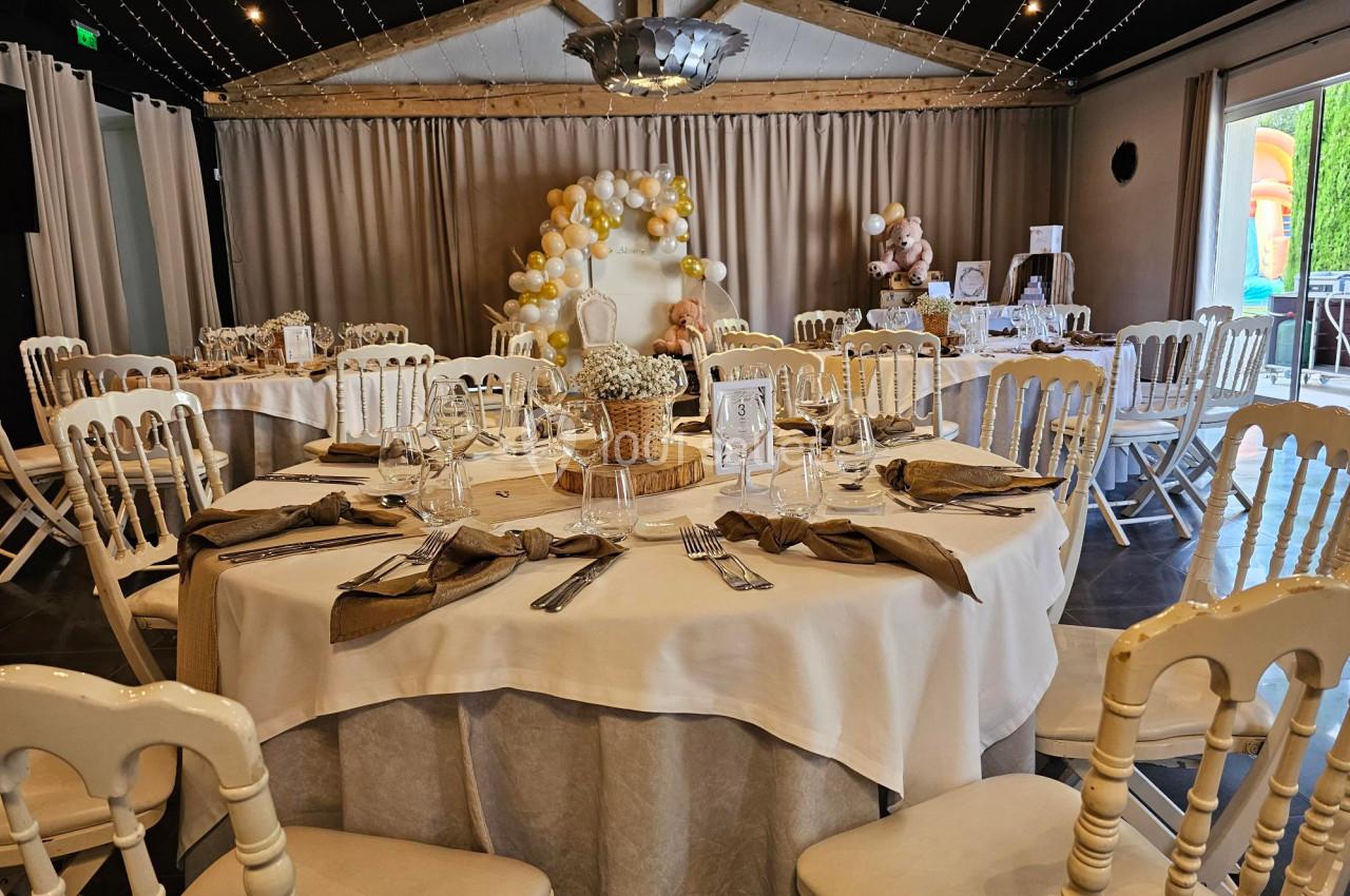 Salle décorée pour un événement avec tables rondes dressées, chaises blanches et arche de ballons en arrière-plan.