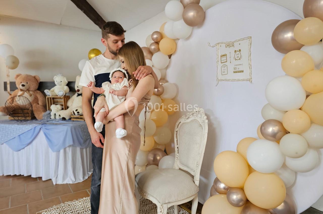 Un couple tenant un bébé dans une salle décorée avec des ballons et des éléments sur le thème de l'enfance.
