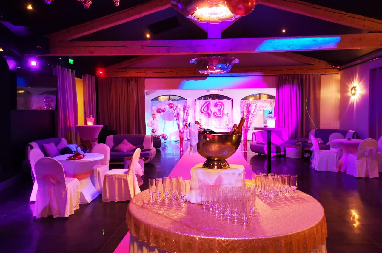 Salle décorée pour une fête avec des tables drapées, des fauteuils, des lumières colorées et une table avec des verres et un…