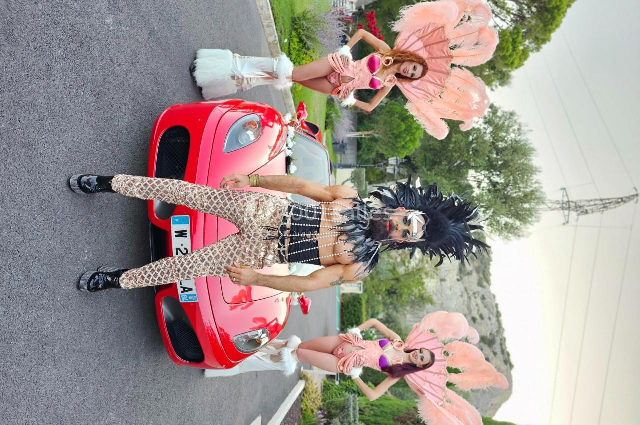 Un homme en tenue extravagante pose devant une voiture rouge, accompagné de deux femmes en costumes à plumes roses.