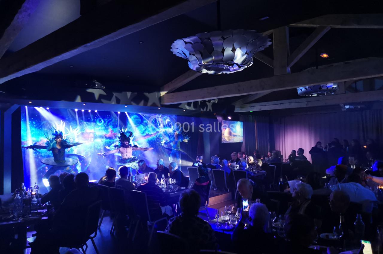 Spectacle de danse avec costumes lumineux sur une scène, devant un public attablé dans une salle sombre.