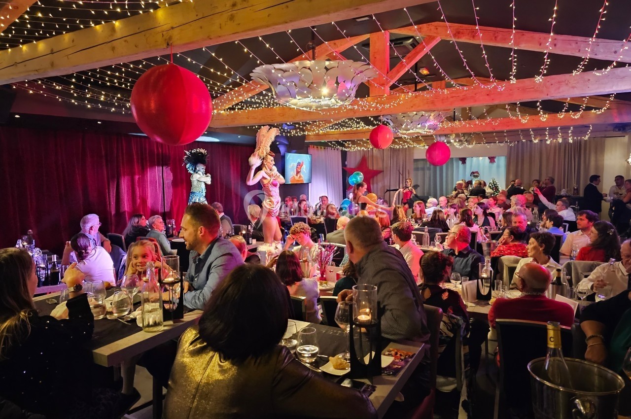 Salle de restaurant animée avec des convives attablés et un spectacle de danseuses sous des guirlandes lumineuses.