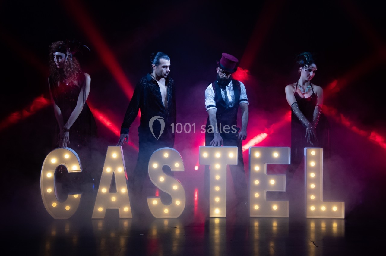 Quatre personnes posent derrière des lettres lumineuses formant ’CASTEL’, avec un éclairage rouge et une ambiance de scène.