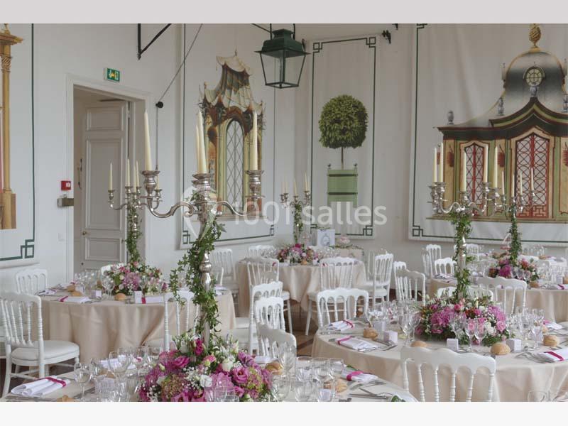 Salle de réception élégante décorée de chandeliers, fleurs roses et tables dressées avec nappes beiges.