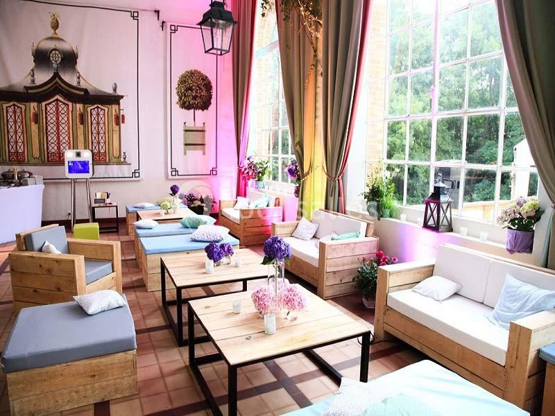 Salon lumineux avec de grandes fenêtres, mobilier en bois, coussins colorés et décorations florales.