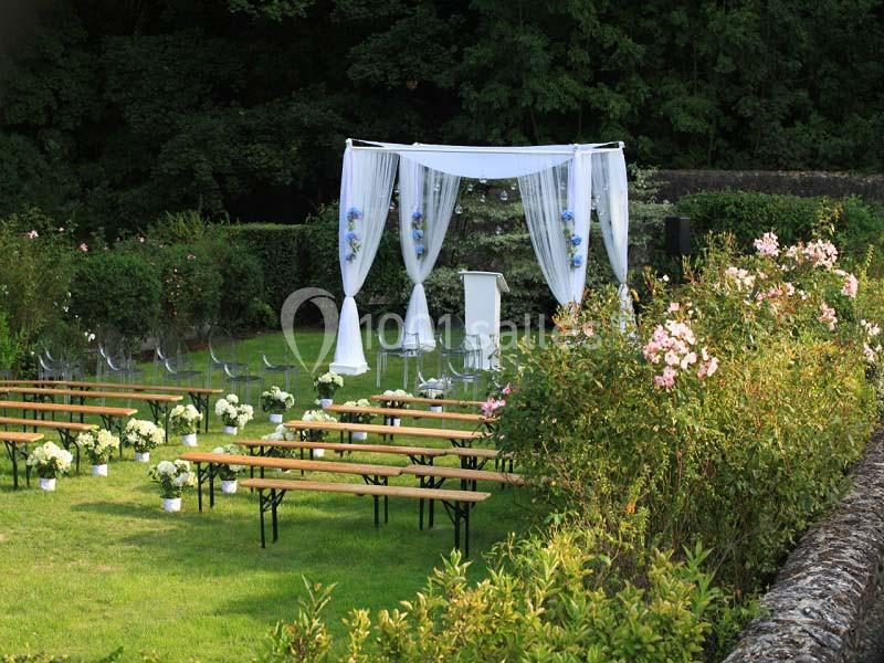 Structure de cérémonie en plein air avec arche blanche décorée de fleurs, bancs en bois et chaises transparentes sur pelouse.