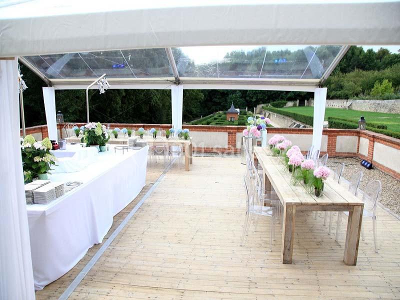 Salle de réception extérieure avec tables en bois décorées de fleurs, sous une structure transparente.