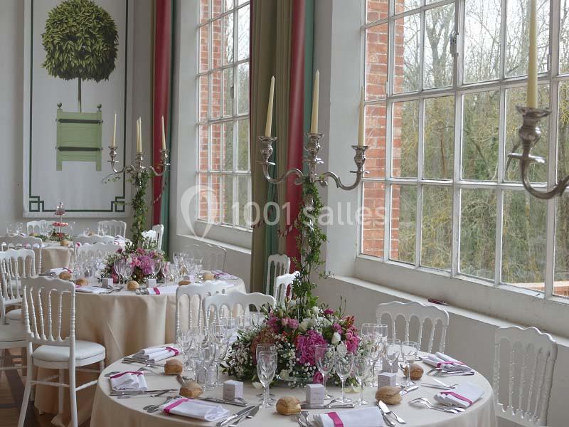 Salle lumineuse avec tables rondes décorées de fleurs, chandeliers et vaisselle élégante, près de grandes fenêtres.