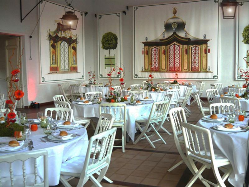 Salle décorée pour un événement avec tables rondes dressées, chaises blanches et fresques murales élégantes.