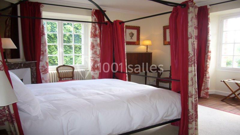 Chambre lumineuse avec lit à baldaquin, rideaux rouges, mobilier en bois et fenêtres donnant sur un jardin verdoyant.