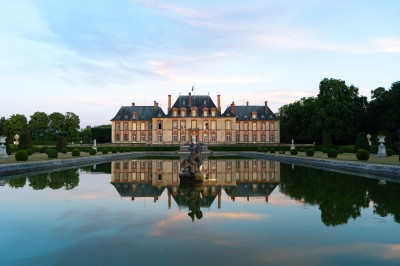 Miniature Le château de Breteuil Salon lumineux avec fauteuils bleus, table basse, cheminée, lampes, cadres au mur et grande fenêtre avec rideaux fleuris.