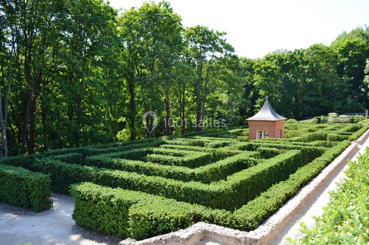 Labyrinthe de haies taillées entouré d'arbres, avec une petite cabane en briques au centre.