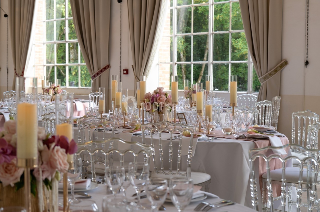 Salle de réception élégante avec tables dressées, décorations florales roses et chandeliers, près de grandes fenêtres.