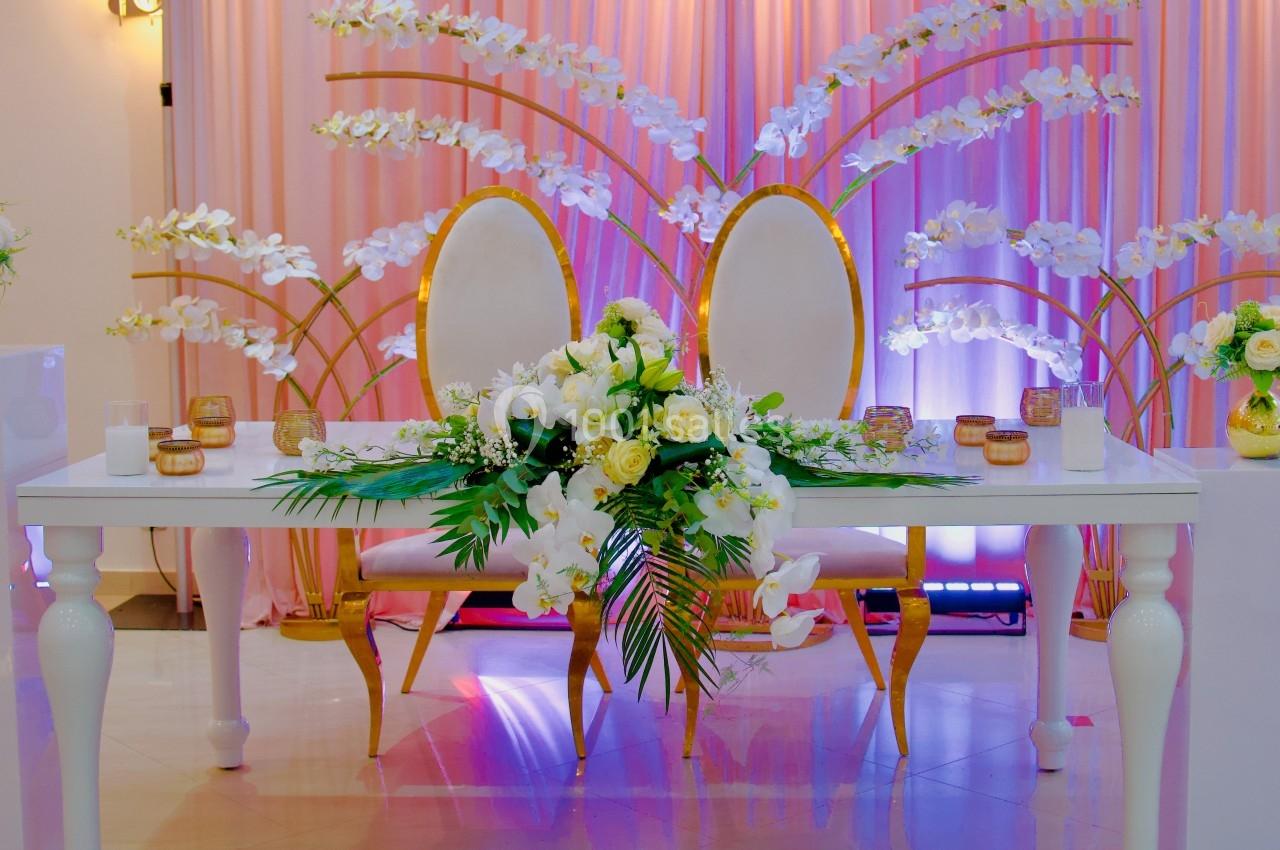 Table décorée avec des fleurs blanches et jaunes, deux chaises élégantes et un fond rose orné d'orchidées.