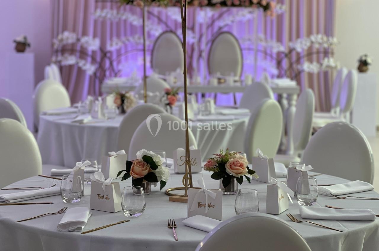 Salle de réception décorée avec des tables rondes, nappes blanches, fleurs et éclairage violet en arrière-plan.