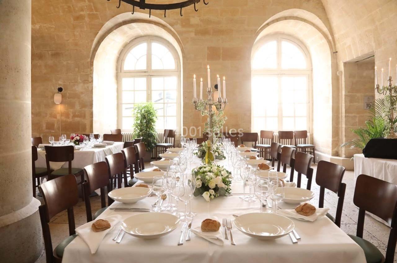 Salle de réception lumineuse avec une grande table dressée, ornée de fleurs et de chandeliers, entourée de chaises.