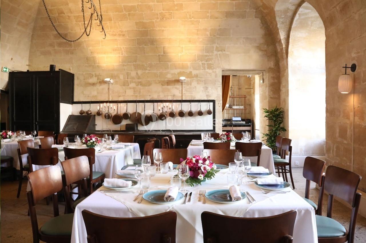 Salle de restaurant élégante avec tables dressées, nappes blanches, chaises en bois et murs en pierre beige.