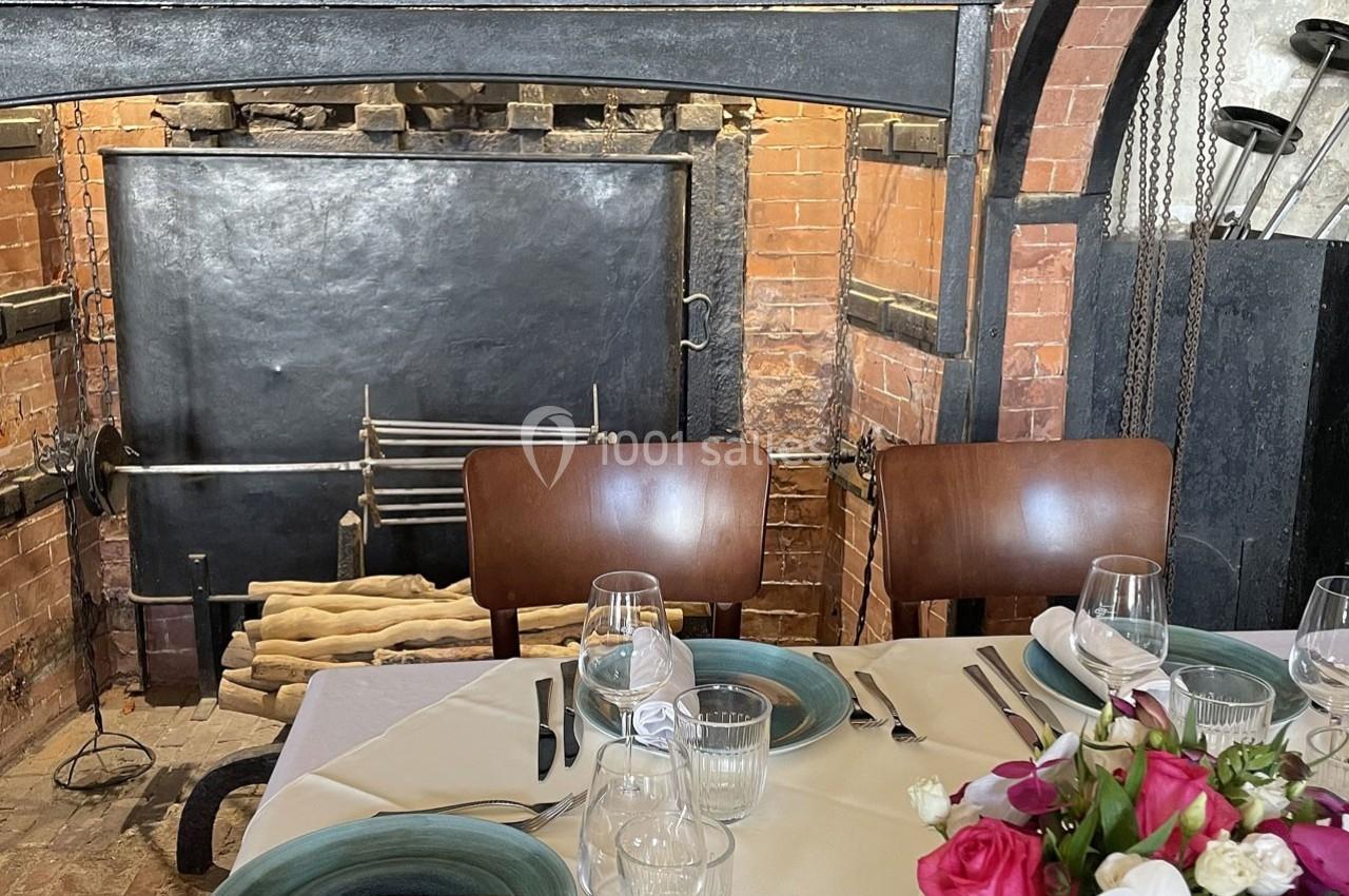 Table dressée avec vaisselle et fleurs devant une cheminée en briques avec des outils de foyer suspendus.