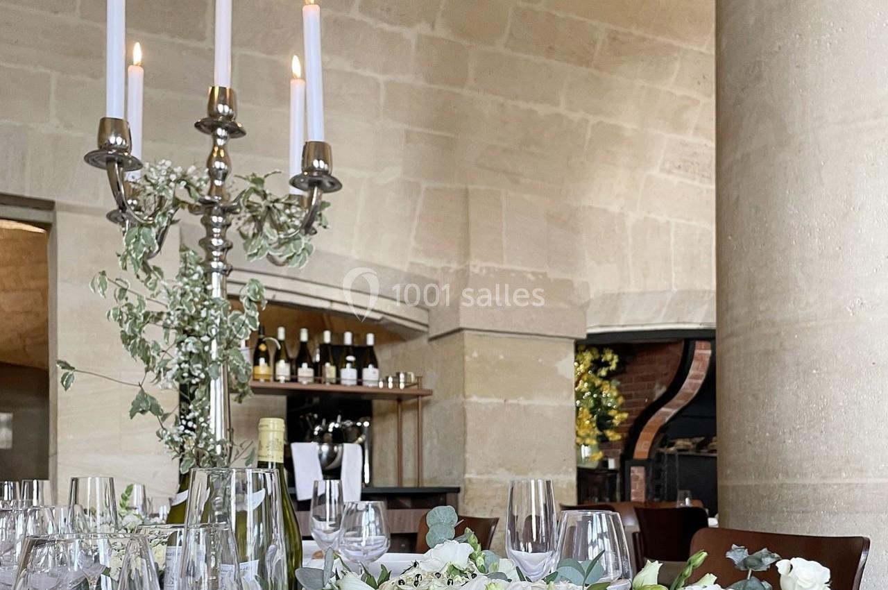 Table élégamment dressée avec chandeliers, fleurs blanches et verres, dans une salle voûtée en pierre.