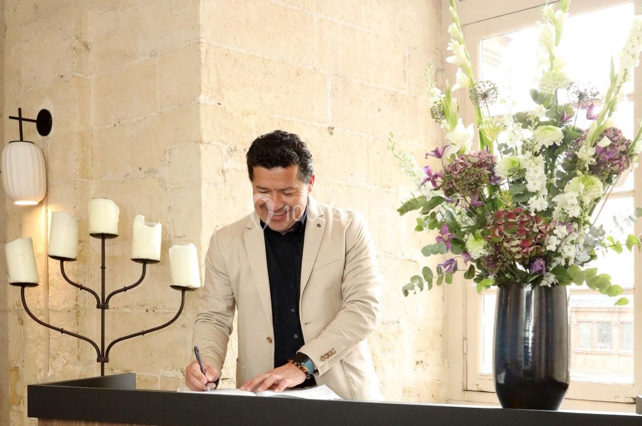 Un homme souriant signe un document sur un comptoir, avec un chandelier et un grand vase de fleurs en arrière-plan.