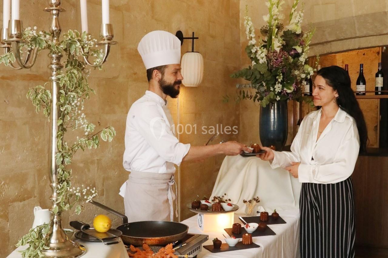 Un chef en tenue blanche sert un dessert à une femme dans un cadre élégant avec des fleurs et des bougies.