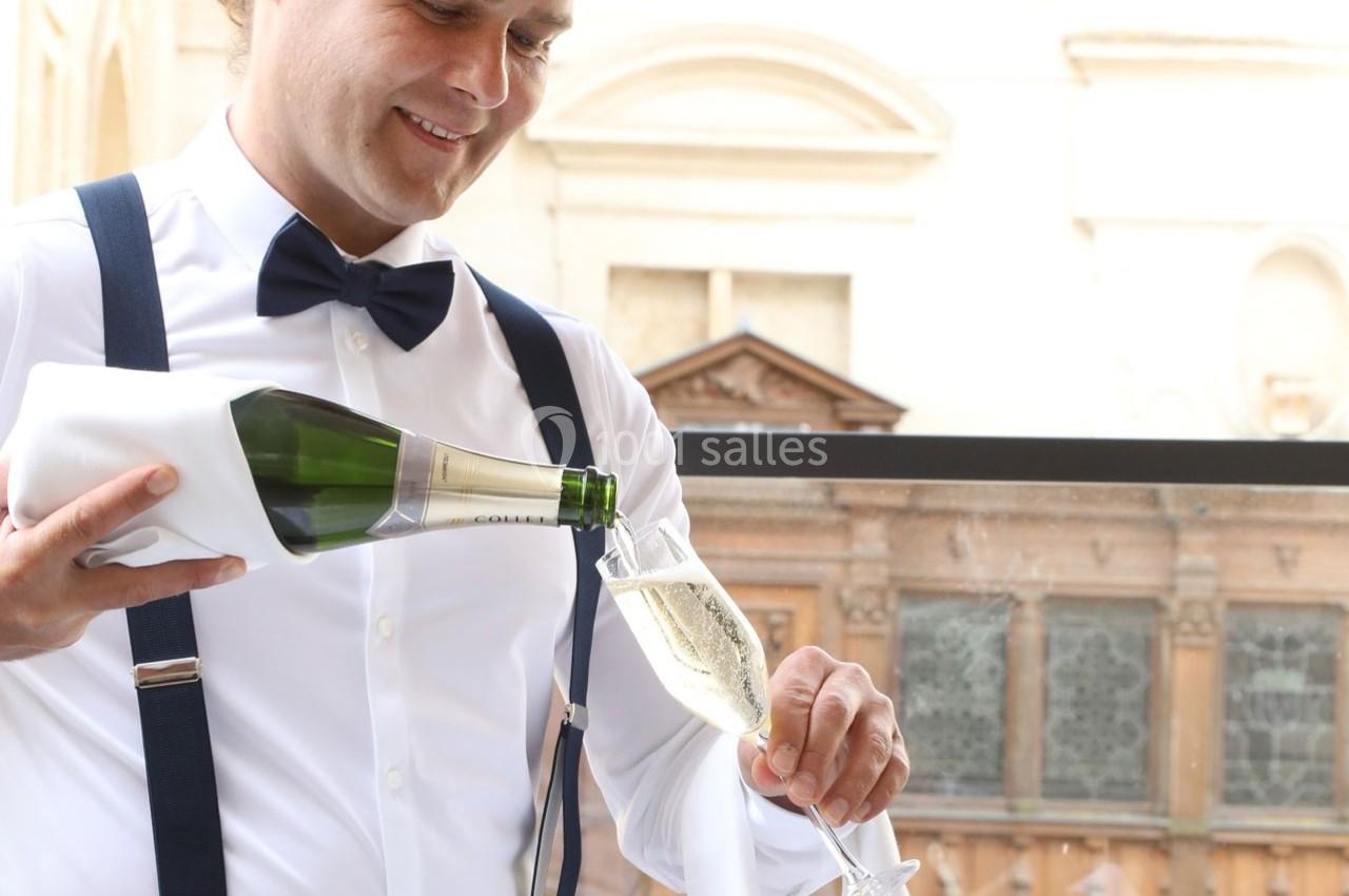 Un serveur en tenue élégante verse du champagne dans une flûte sur une terrasse avec un bâtiment en arrière-plan.