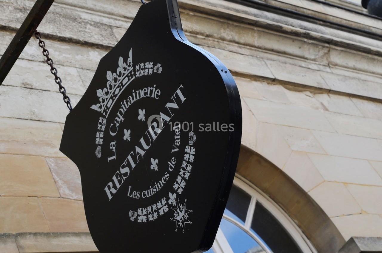 Enseigne noire suspendue indiquant ’La Capitainerie Restaurant, Les cuisines de Vatel’ devant un bâtiment en pierre.
