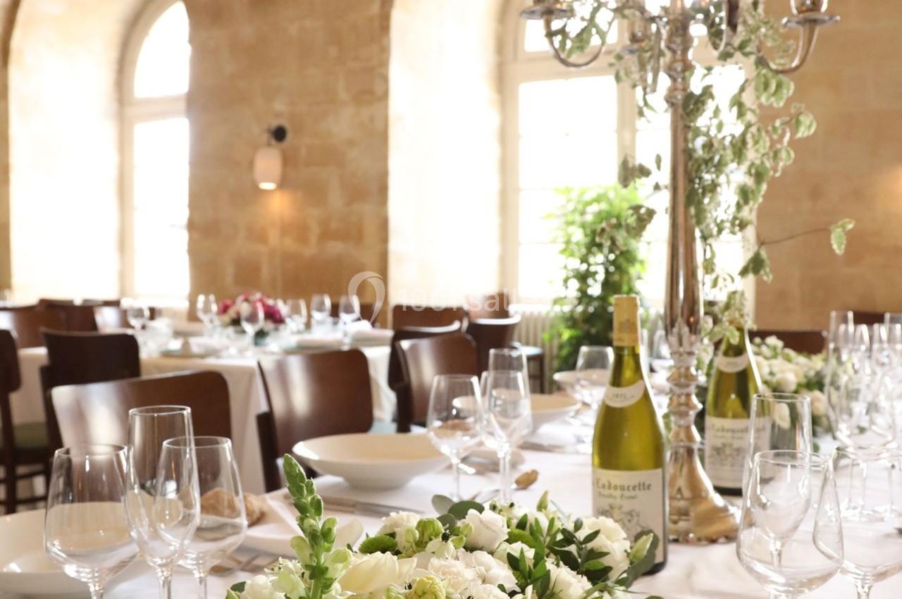 Salle de réception lumineuse avec tables dressées, décorations florales et bouteilles de vin.
