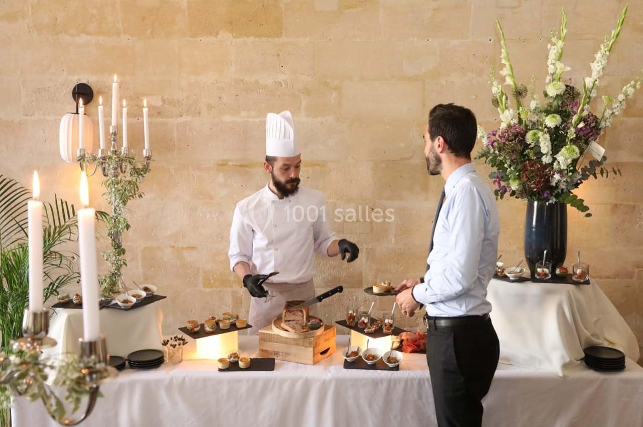 Un chef en tenue blanche prépare des plats devant un invité à une table garnie de mets et décorée de fleurs.
