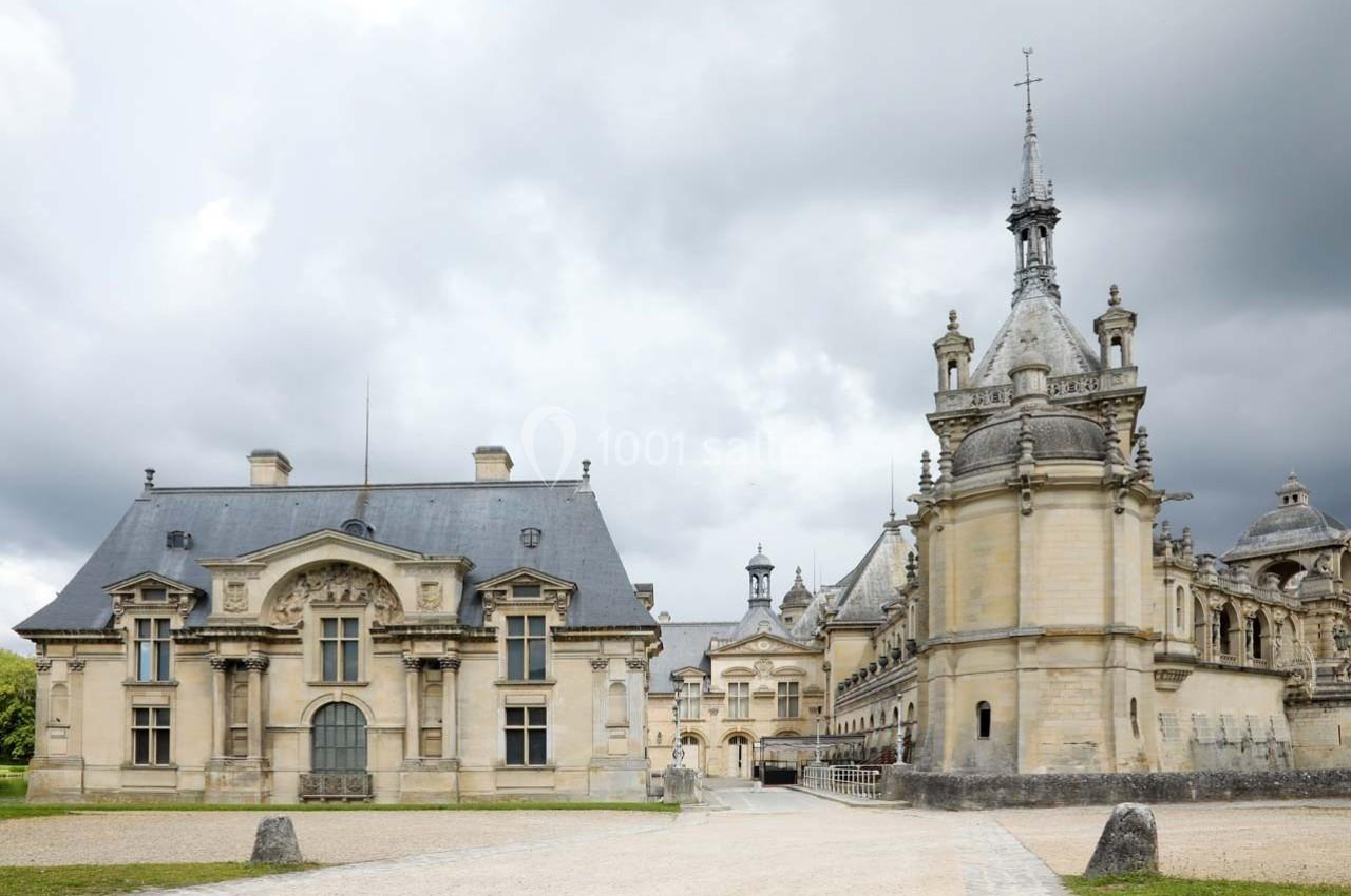 Façade d'un château historique avec des bâtiments en pierre, toits en ardoise et ciel nuageux en arrière-plan.