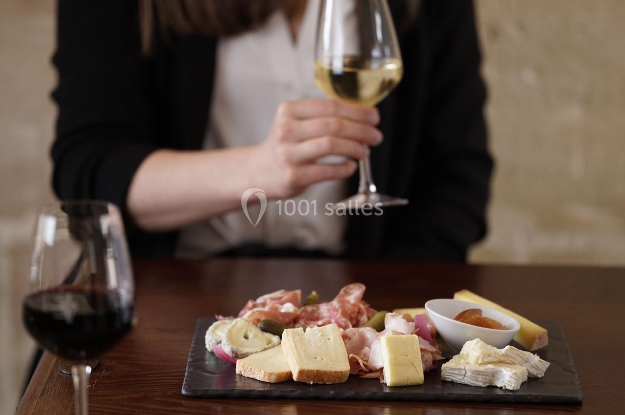 Planche de fromages et charcuteries sur une table, avec une personne tenant un verre de vin blanc en arrière-plan.