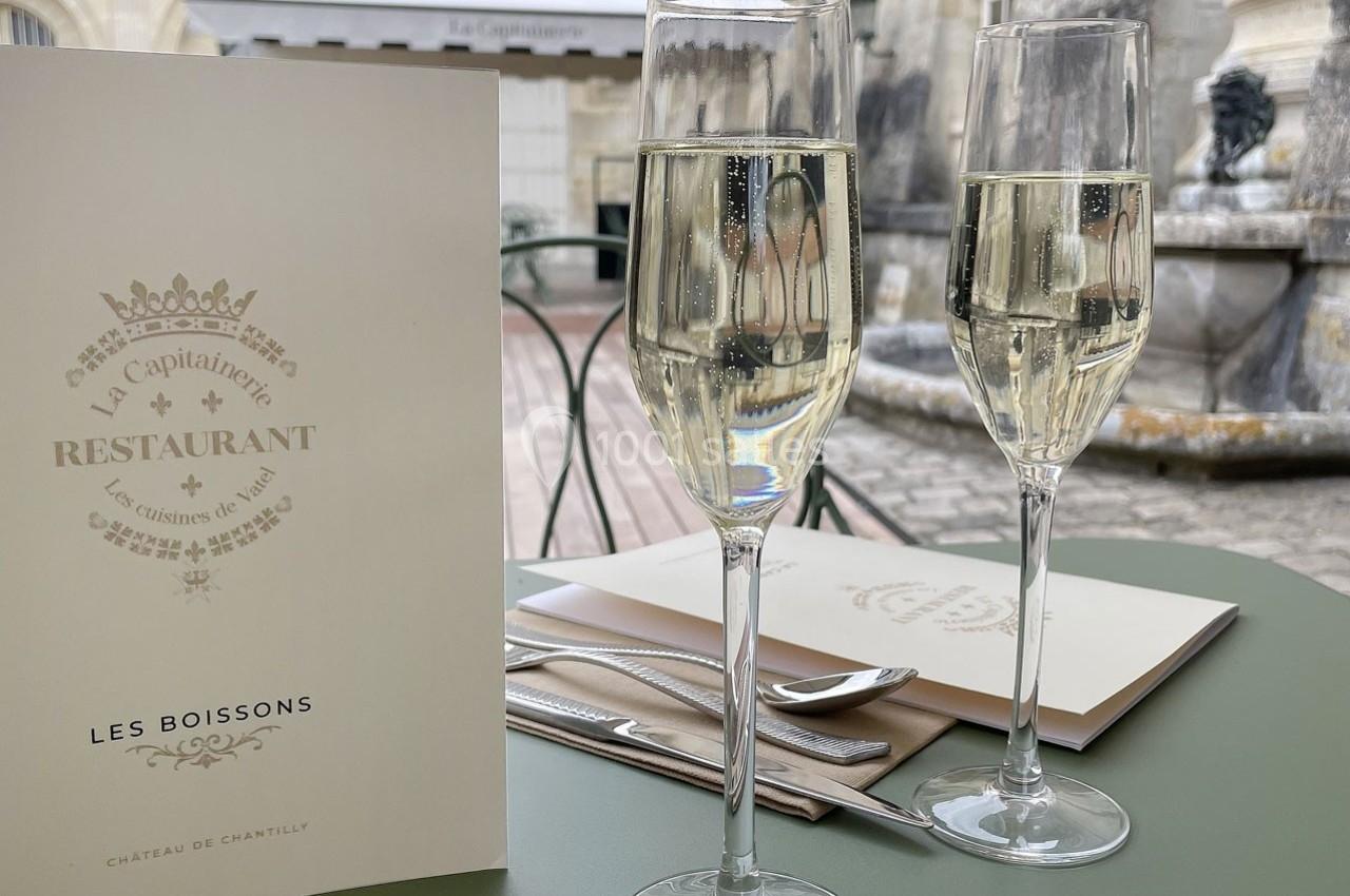 Deux verres de champagne sur une table de restaurant en terrasse, avec un menu et un décor en pierre en arrière-plan.