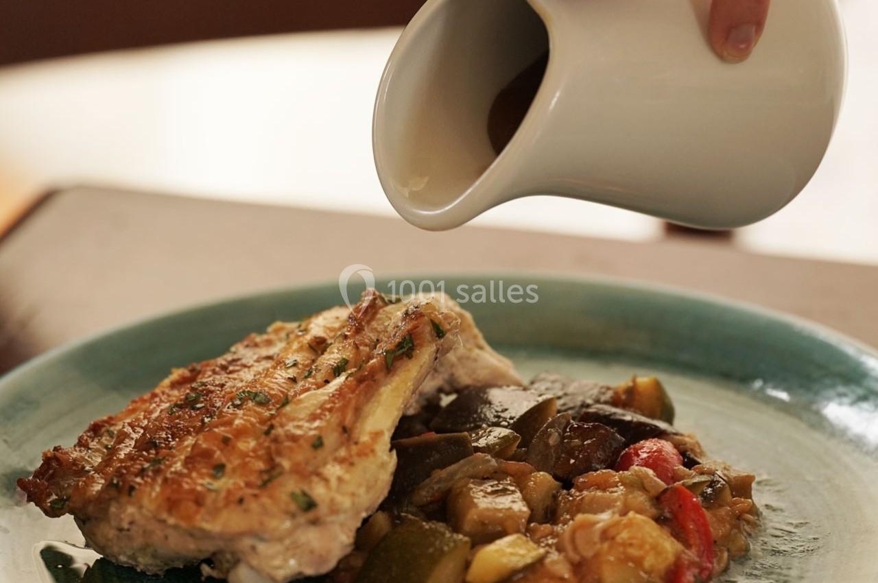 Une main verse une sauce brune depuis un petit pichet sur un plat de poulet grillé accompagné de légumes rôtis.