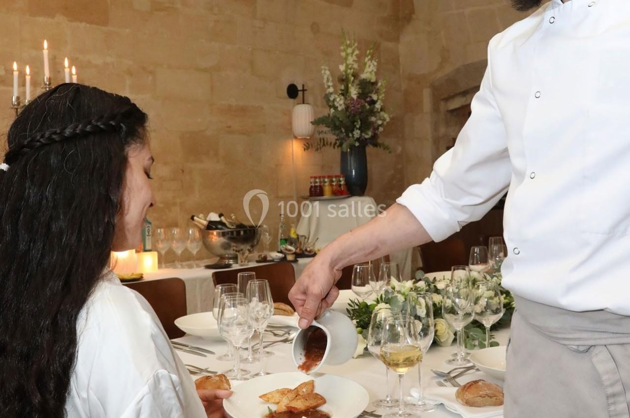 Un chef verse une sauce dans une assiette devant une femme assise à une table élégamment dressée.