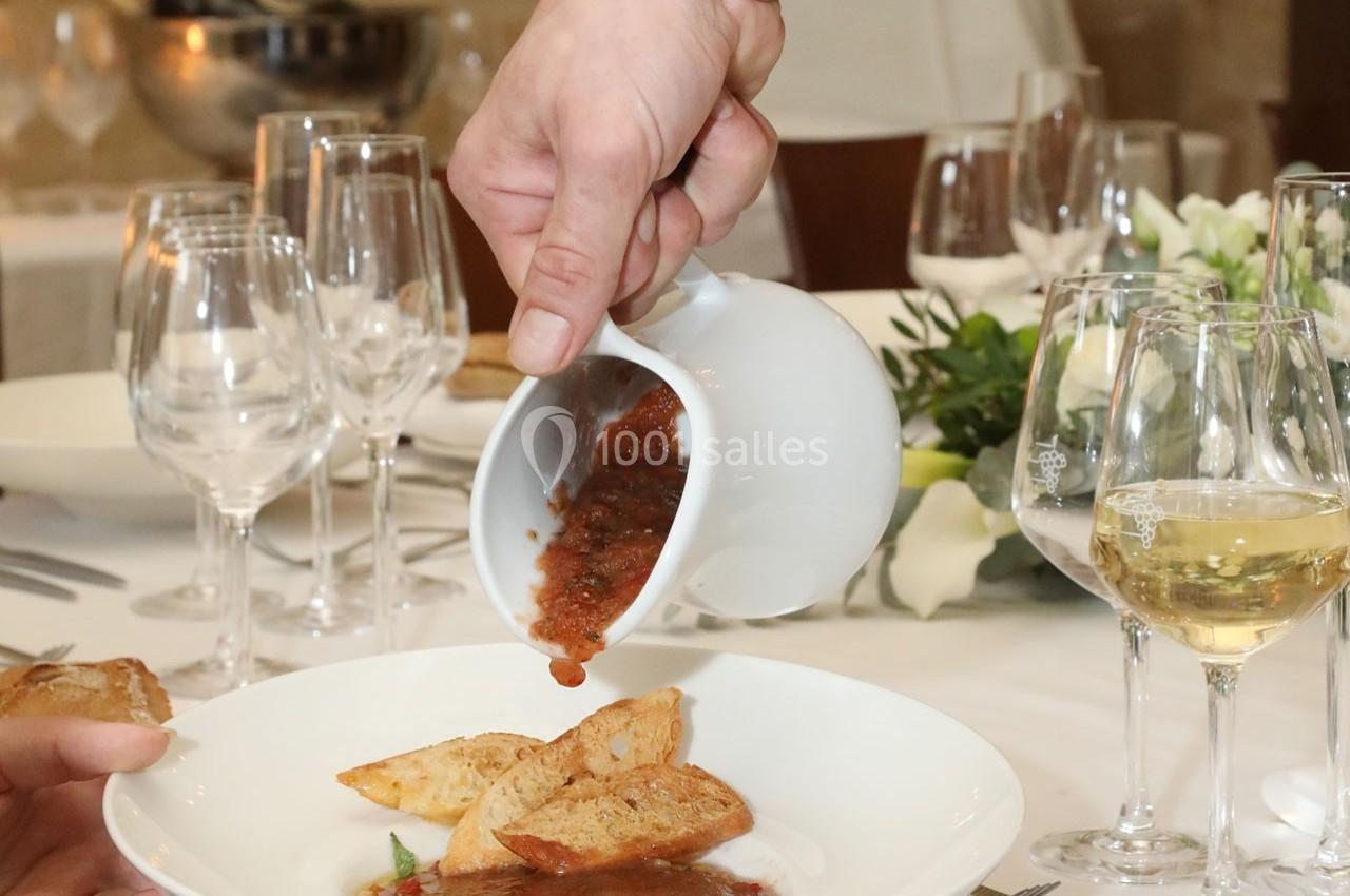 Une main verse une sauce tomate sur des morceaux de pain dans une assiette, sur une table dressée élégamment.