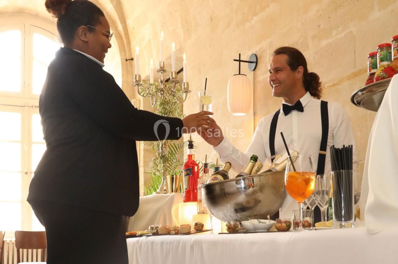 Une femme reçoit un verre de cocktail d'un serveur souriant derrière un bar dans un cadre lumineux et élégant.