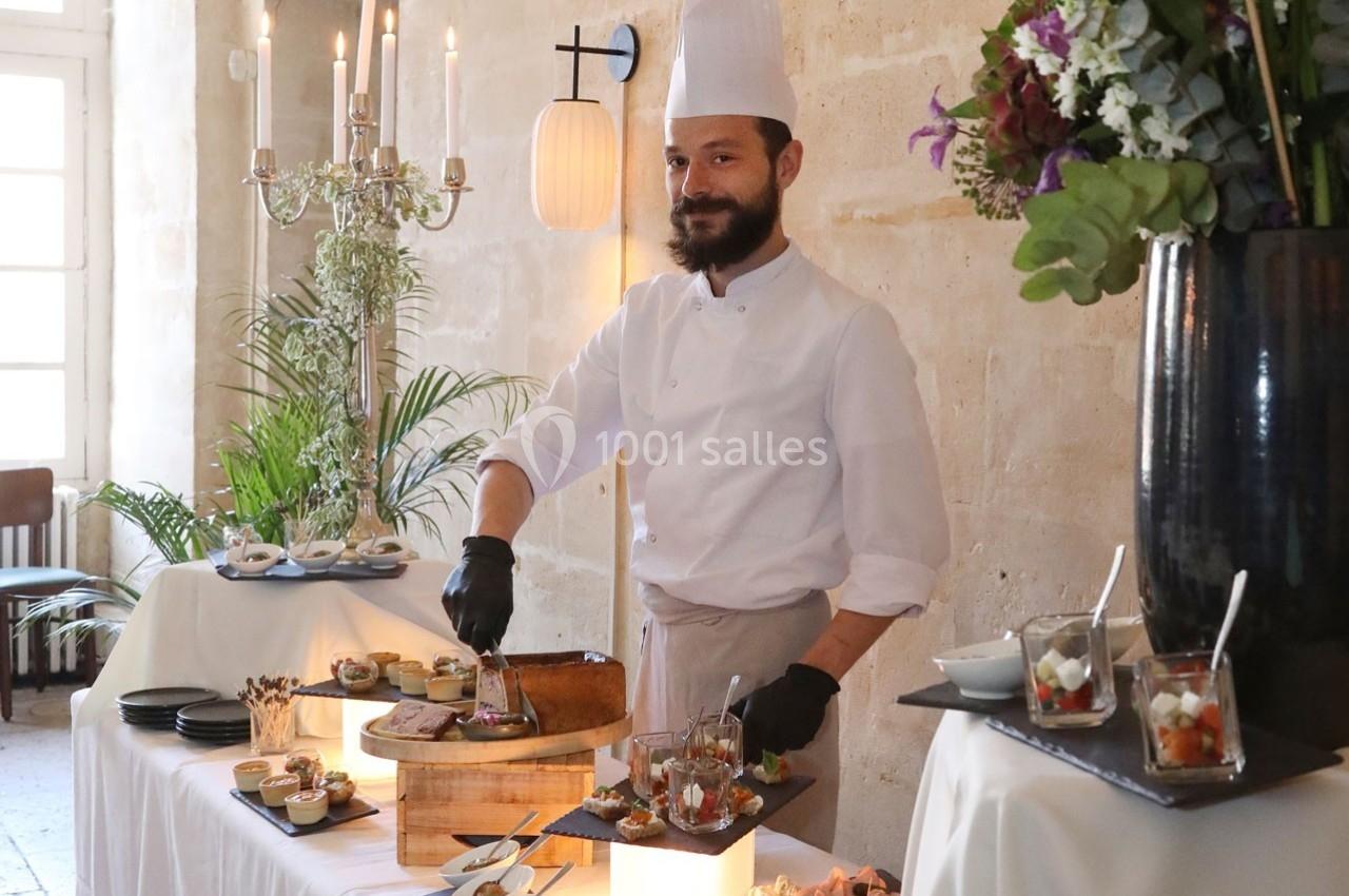 Un chef en tenue blanche prépare des plats sur un buffet décoré, dans une salle lumineuse avec des fleurs et des chandeliers.
