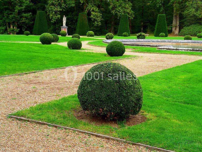 Allée de gravier bordée de pelouses et de buis taillés en boules dans un jardin paysager avec une statue en arrière-plan.